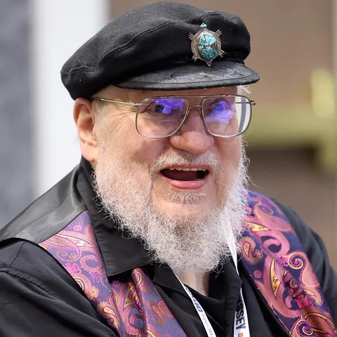 Foto do escritor George R. R. Martin sorrindo, de boina preta e óculos