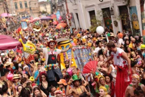 Multidão animada celebrando o Carnaval em uma rua histórica, com pessoas fantasiadas, sombrinhas coloridas, adereços vibrantes e clima de festa ao ar livre.